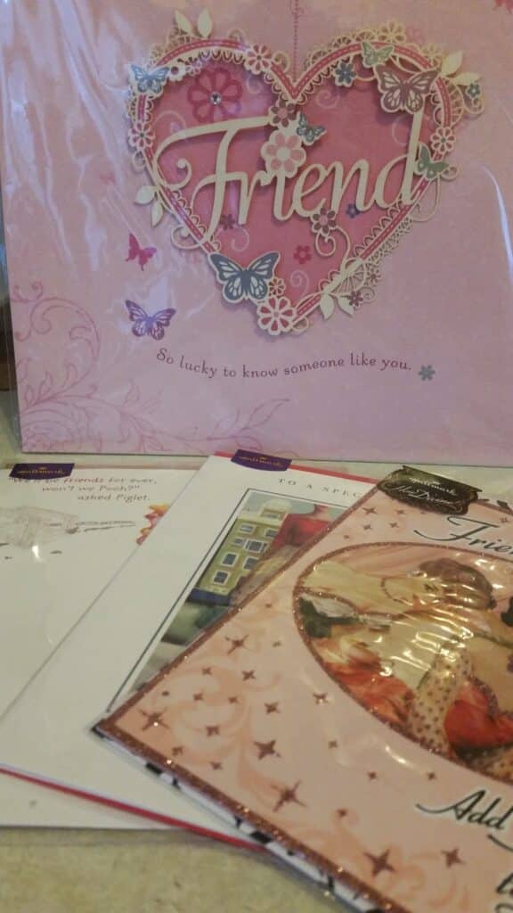 A close-up of several greeting cards, with the largest featuring a pink heart, butterflies, and the word "Friend" in elegant script. Below it reads, "So lucky to know someone like you." Other colourful cards are partially visible in the foreground.