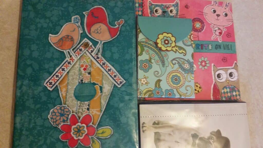 A collection of colourful notebooks and folders with whimsical designs, including birds on a birdhouse, an owl, floral patterns, a cartoon cat, and a pink cover with "WRITE on ME!" text. The bottom right shows a black-and-white image of dogs.