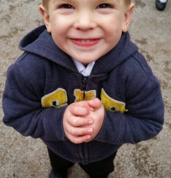 A young child with short blond hair smiles brightly at the camera, standing outside on a gravel surface. The child wears a dark blue hoodie with yellow letters, black trousers, and black shoes, with hands clasped together in front. Another child’s feet are visible in the background.