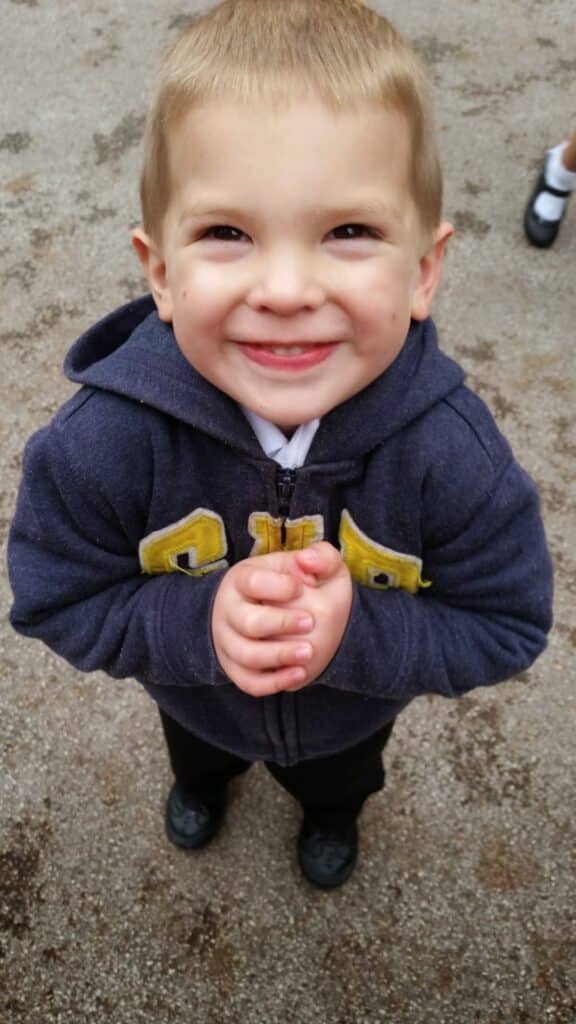 A young child with short light brown hair smiles up at the camera, hands clasped in front. He wears a dark blue zip-up hoodie with yellow letters, dark trousers, and black shoes. The background is a rough, light brown pavement.