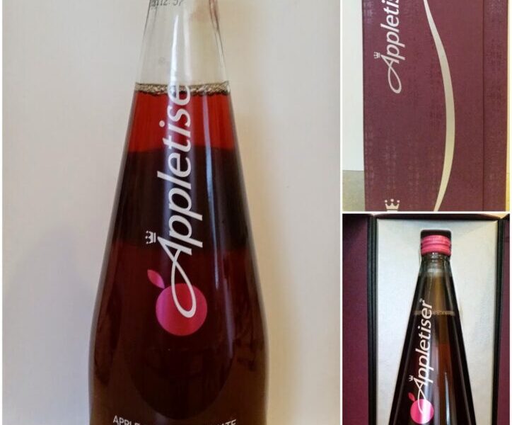 A tall glass bottle of Appletiser Apple & Pomegranate juice with a silver cap, red label, and white text stands on a light surface. Adjacent is a burgundy gift box, open to reveal the bottle inside, both displaying the Appletiser logo.