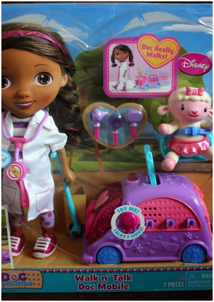 A boxed Doc McStuffins “Walk n’ Talk Doc Mobile” toy set, featuring a Doc McStuffins doll in a white coat, a pink and purple mobile car, a figure of Lambie, medical tools, and bright, colourful packaging with Disney and Doc McStuffins logos.