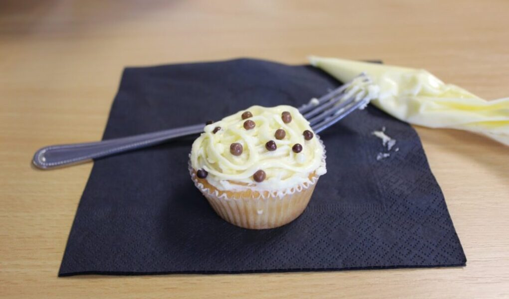 A vanilla cupcake with white and yellow swirled icing and small chocolate balls sits on a black serviette. A piping bag and a fork, both with some leftover icing, are placed beside the cupcake on a light wooden table.