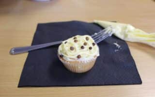 A vanilla cupcake with white and yellow swirled icing and small chocolate balls sits on a black serviette. A piping bag and a fork, both with some leftover icing, are placed beside the cupcake on a light wooden table.