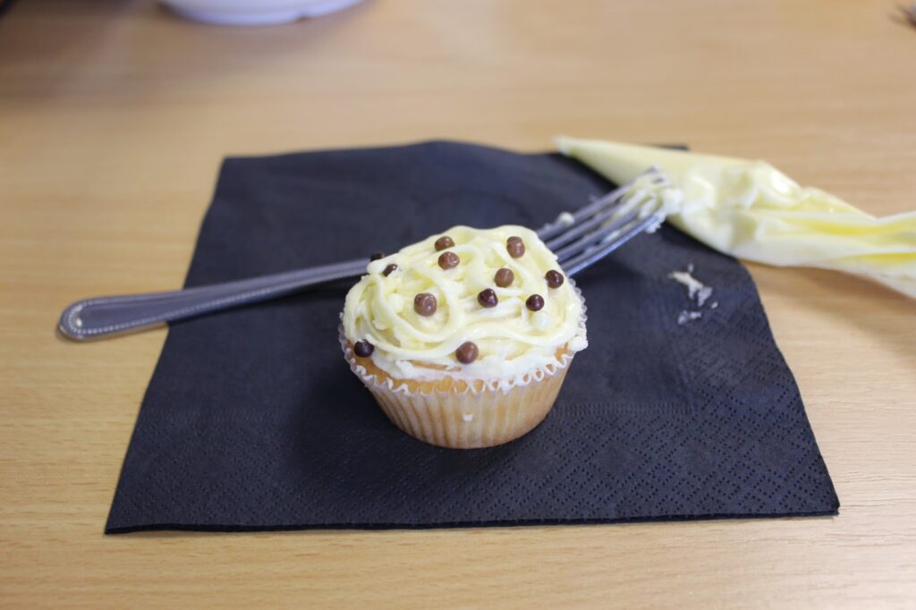A vanilla cupcake with yellow icing and small chocolate balls sits on a black serviette. A metal fork and an empty piping bag are in the background, all placed on a light wooden surface.