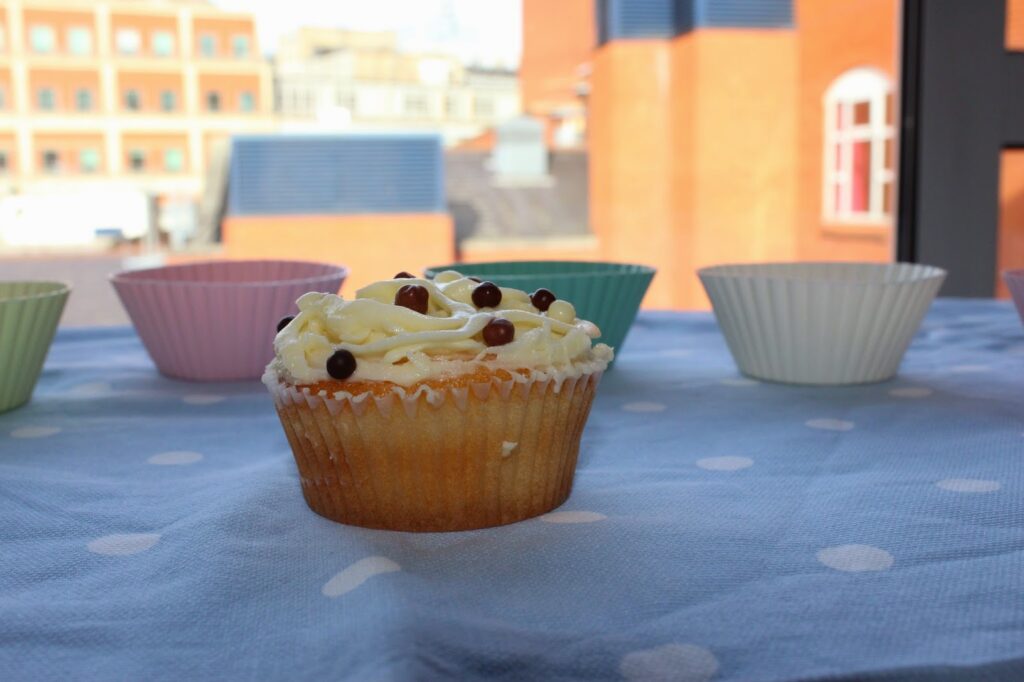 A vanilla cupcake with white icing and chocolate pearls sits on a blue polka dot tablecloth. Empty, colourful cupcake cases are in the background, and orange brick buildings are visible through a window behind the table.