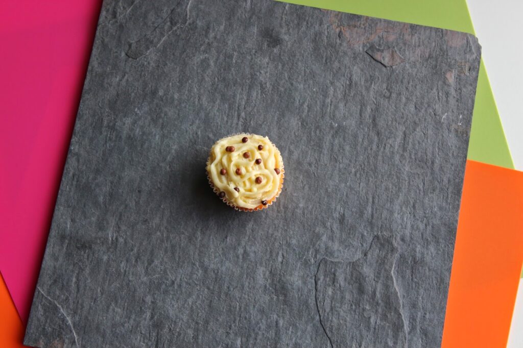 A single cupcake with swirled white icing and brown sprinkles sits centred on a dark grey textured slate. The slate is laid atop bright pink, lime green, and orange paper sheets, their edges visible in the corners of the image.
