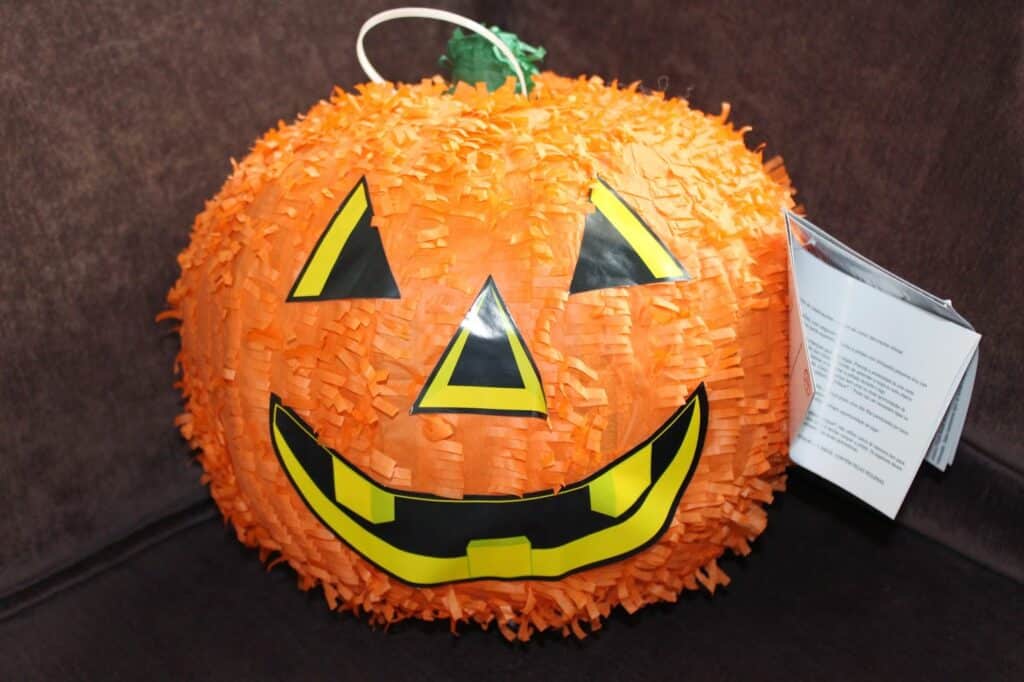 A round pumpkin piñata with orange fringed paper, a green stalk, and a smiling jack-o’-lantern face made of black and yellow paper. The piñata has a white hanging string and a label attached to its side. The background is a dark brown fabric.