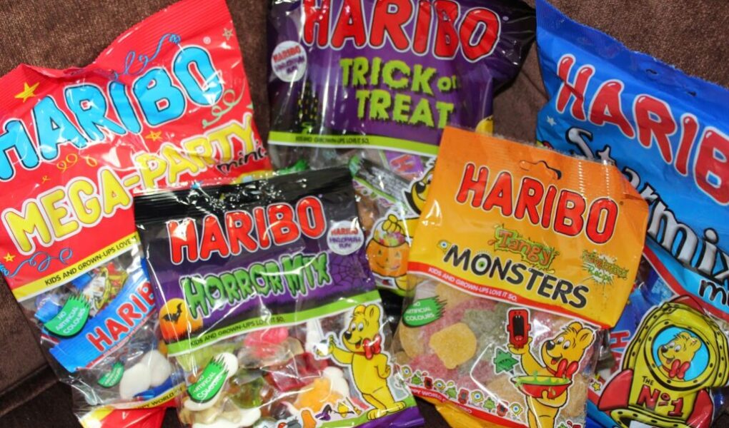 Several colourful bags of HARIBO gummy sweets are displayed on a brown surface. Visible flavours include Mega Party, Trick or Treat, Starmix, HorrorMix, and Terrifying Monsters, each bag featuring playful designs and cartoon gummy bears.