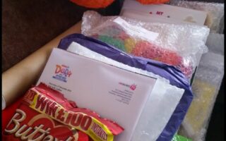 A colourful flower-shaped piñata sits above a stack of bubble-wrapped packages, papers, a rolled poster, a red bag of Butterkist Toffee Popcorn, and several green containers with purple cartoon monkeys, all arranged on a flat surface.