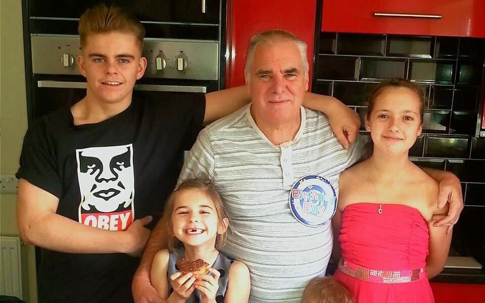 A smiling older man with grey hair stands in a kitchen with three children and a teenager. The man wears a "Birthday Boy" badge. The girl in a red dress and the older boy in an "Obey" shirt stand on either side, while two younger children hold cupcakes and smile.