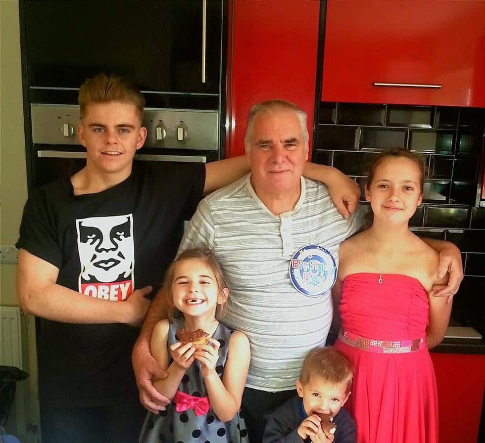 A smiling older man with grey hair stands in a kitchen with three children and a teenager. The man wears a "Birthday Boy" badge. The girl in a red dress and the older boy in an "Obey" shirt stand on either side, while two younger children hold cupcakes and smile.