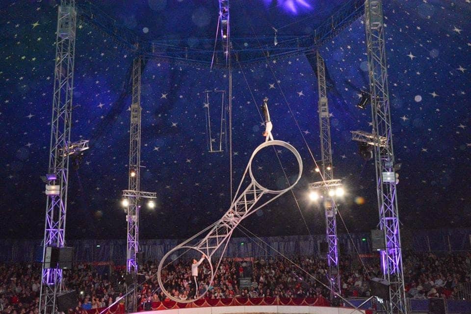 A circus performer stands on top of a large rotating wheel apparatus, balancing on one hand high above the audience. The circus tent is dark blue with white stars, and spotlights illuminate the performers while a crowd watches below.
