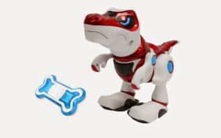 A toy robot dinosaur with red, white, and blue accents stands on a white background. It has large eyes and a friendly expression. Beside it lies a blue and white bone-shaped remote control.
