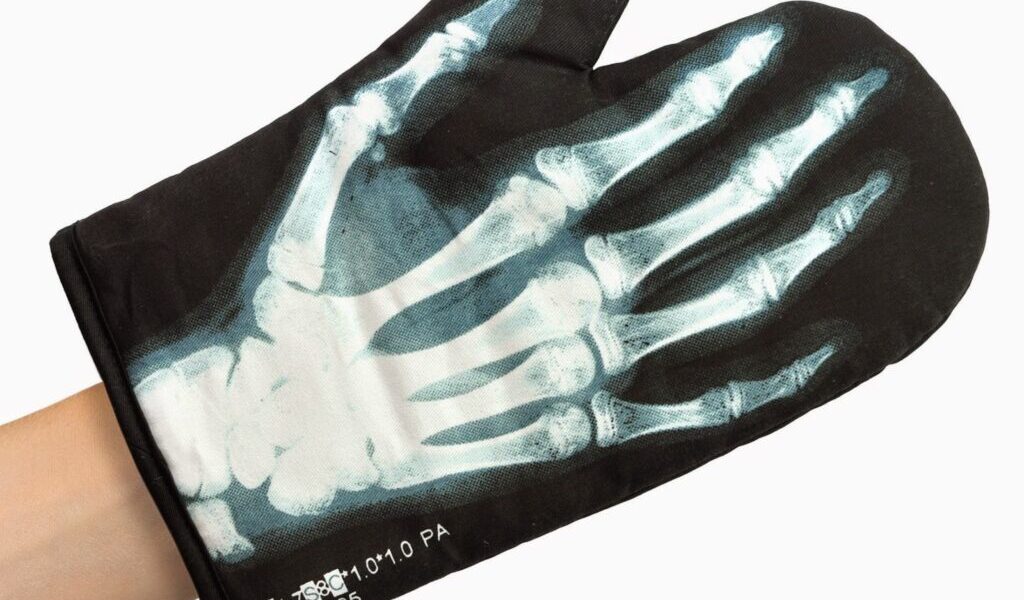 A black oven glove features a printed X-ray image of a human hand, showing white bones and joints in detail. The glove is worn on a left hand, with a small hanging loop visible, and the background is plain white.