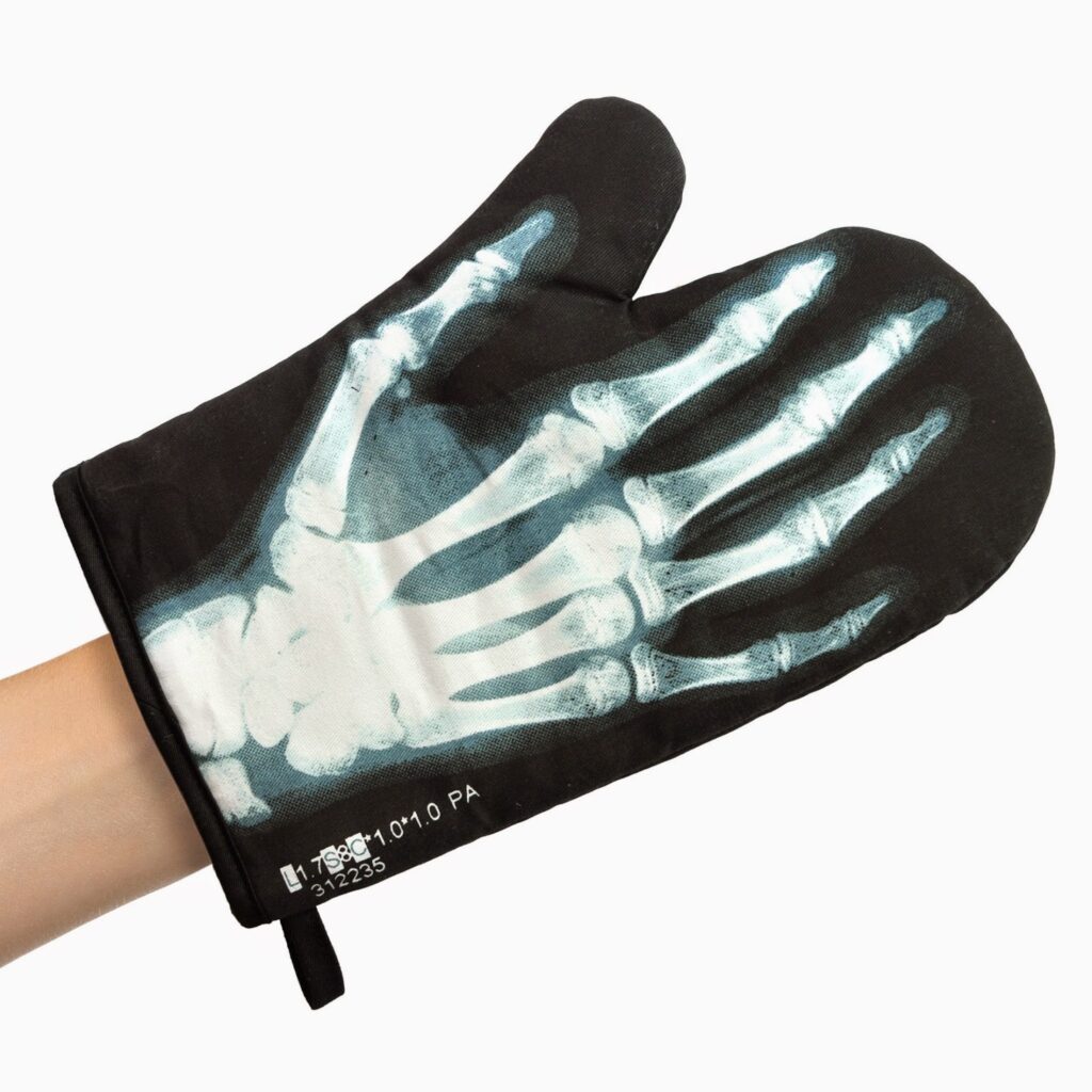 A person’s hand wears a black oven glove printed with an X-ray image of bones, aligned so the fake bones line up with the real hand inside. The glove covers the hand and wrist, with a small hanging loop on the cuff, against a plain white background.