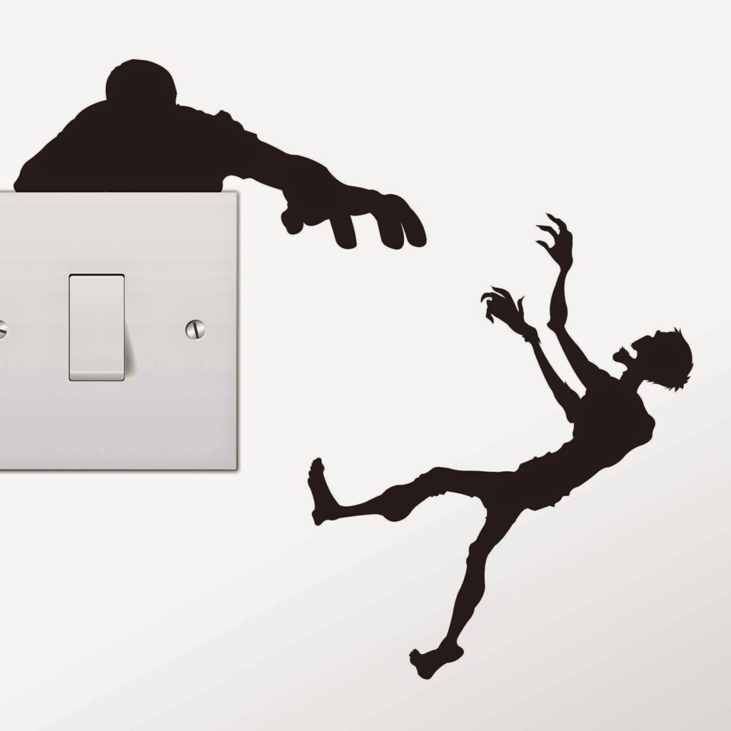 A light switch features black silhouette stickers: a zombie hand emerges from above the switch, reaching towards a zombie figure on the wall. The zombie appears startled, leaning back with outstretched arms, creating a playful, spooky scene.