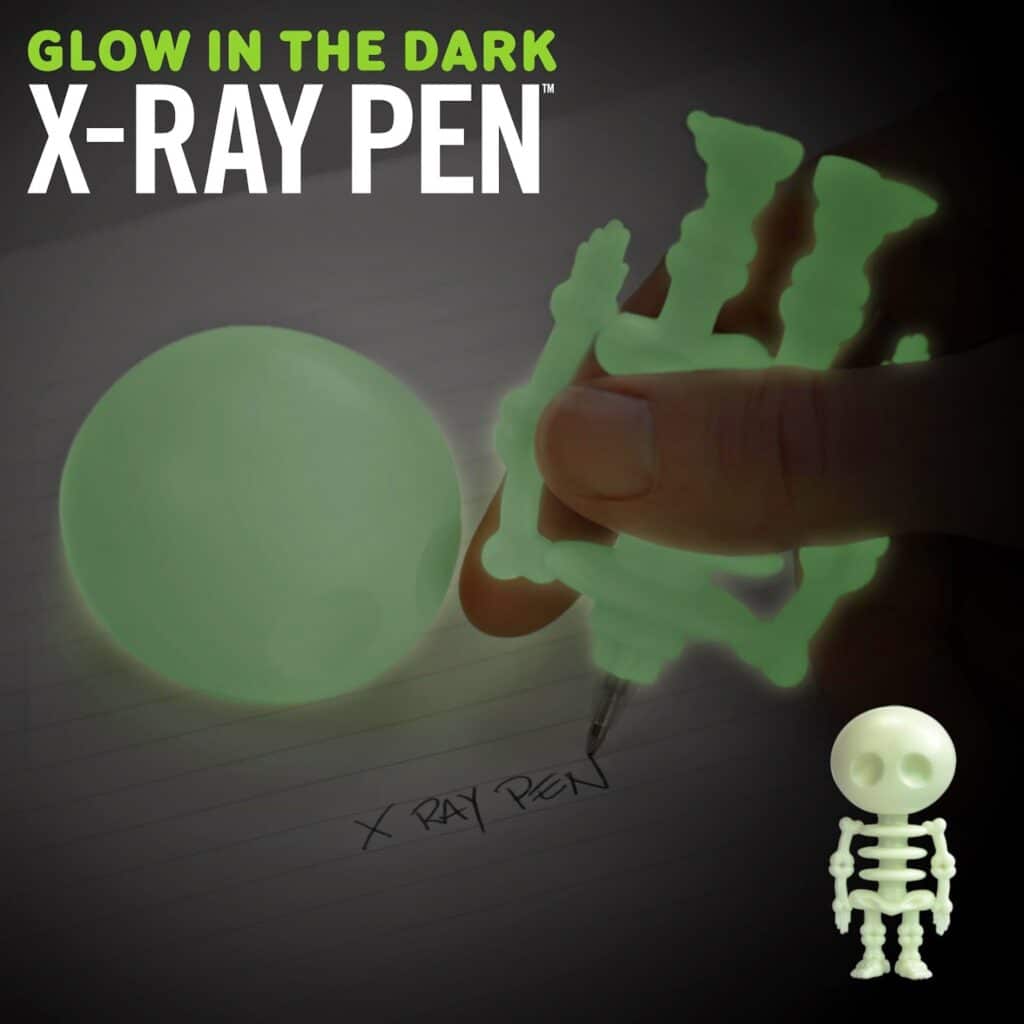 A hand holds a glow-in-the-dark pen shaped like a skeleton, writing “X-RAY PEN” on paper. A glowing green ball and a small glowing skeleton figure are also visible. Text at the top reads, "GLOW IN THE DARK X-RAY PEN.