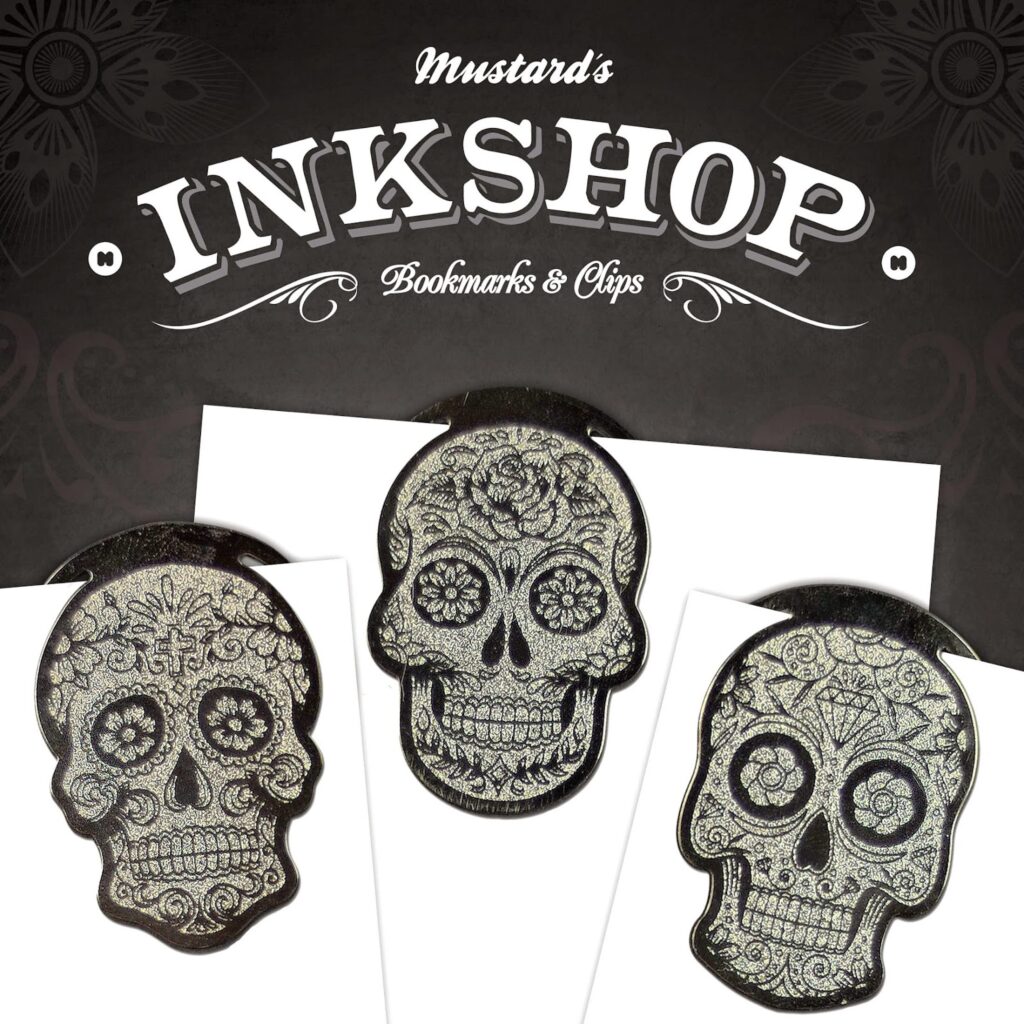 Three metallic skull-shaped bookmarks with intricate, floral Day of the Dead-style designs holding the tops of white cards. Above them, ornate text reads “Mustard’s Inkshop Bookmarks & Clips” on a dark, patterned background.