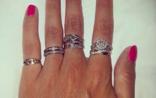A left hand with bright pink nail varnish is shown against a white background. The hand is wearing five ornate silver rings: one on each finger except the thumb, with intricate designs and one featuring a cluster of small stones on the ring finger.