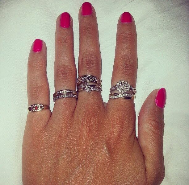A left hand with bright pink nail varnish is shown against a white background. The hand is wearing five ornate silver rings: one on each finger except the thumb, with intricate designs and one featuring a cluster of small stones on the ring finger.