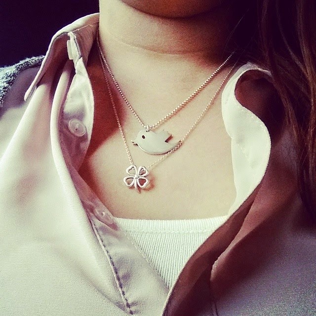 A close-up of a person wearing a pale pink button-up shirt with the top buttons undone, revealing two necklaces: one with a silver bird pendant and another with a silver four-leaf clover pendant, over a white camisole underneath.