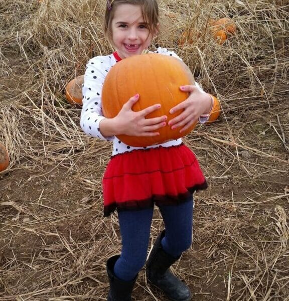 A smiling young girl stands in a pumpkin patch, holding a large orange pumpkin. She wears a white polka dot top, red skirt, blue leggings, and black boots. The ground is dry with straw and scattered pumpkins; other people are visible in the background.