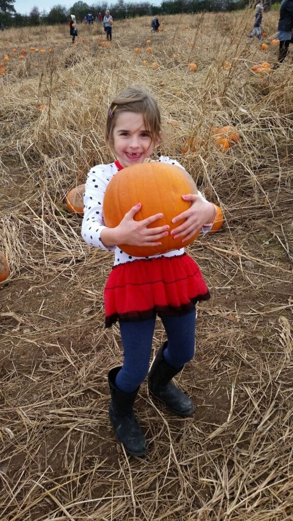 A young girl in a white shirt with black polka dots, a red skirt, blue leggings, and black boots stands in a pumpkin patch, smiling and holding a large orange pumpkin. Dry grass and scattered pumpkins cover the ground; people are in the background.