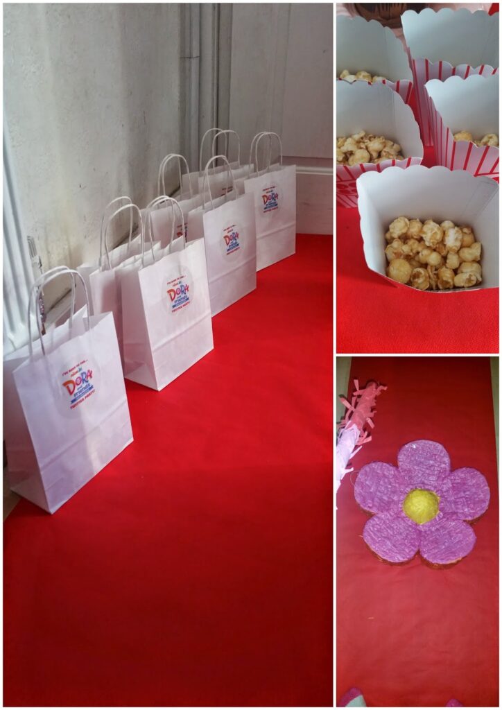 A collage shows: left, five white gift bags with labels on a red tablecloth; top right, popcorn in striped cups; bottom right, a large, textured purple flower with a yellow centre, likely a decoration, on the same red surface.