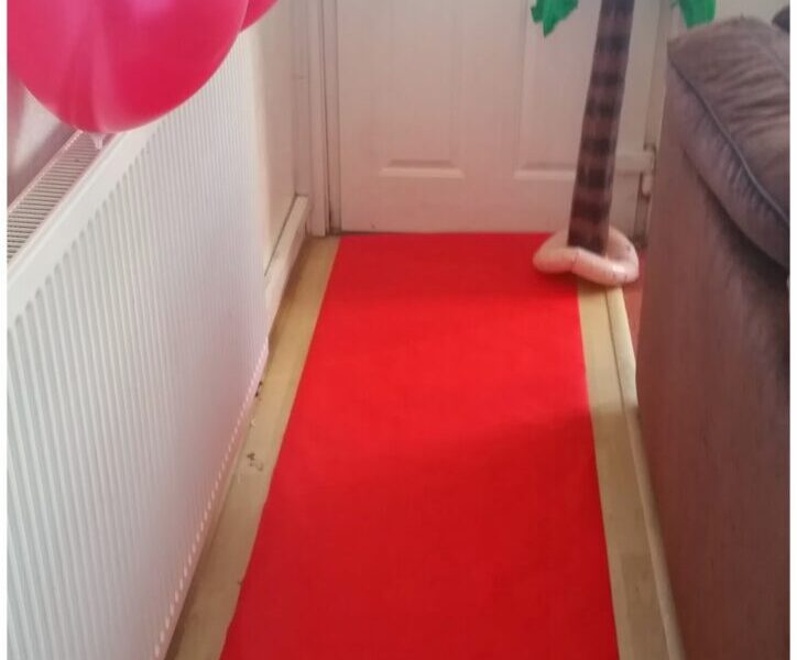 A red carpet runs from the foreground to a white door. Three red balloons are attached to a radiator on the left, and an inflatable palm tree stands by the door on the right, next to a brown sofa. A green object hangs from the door handle.