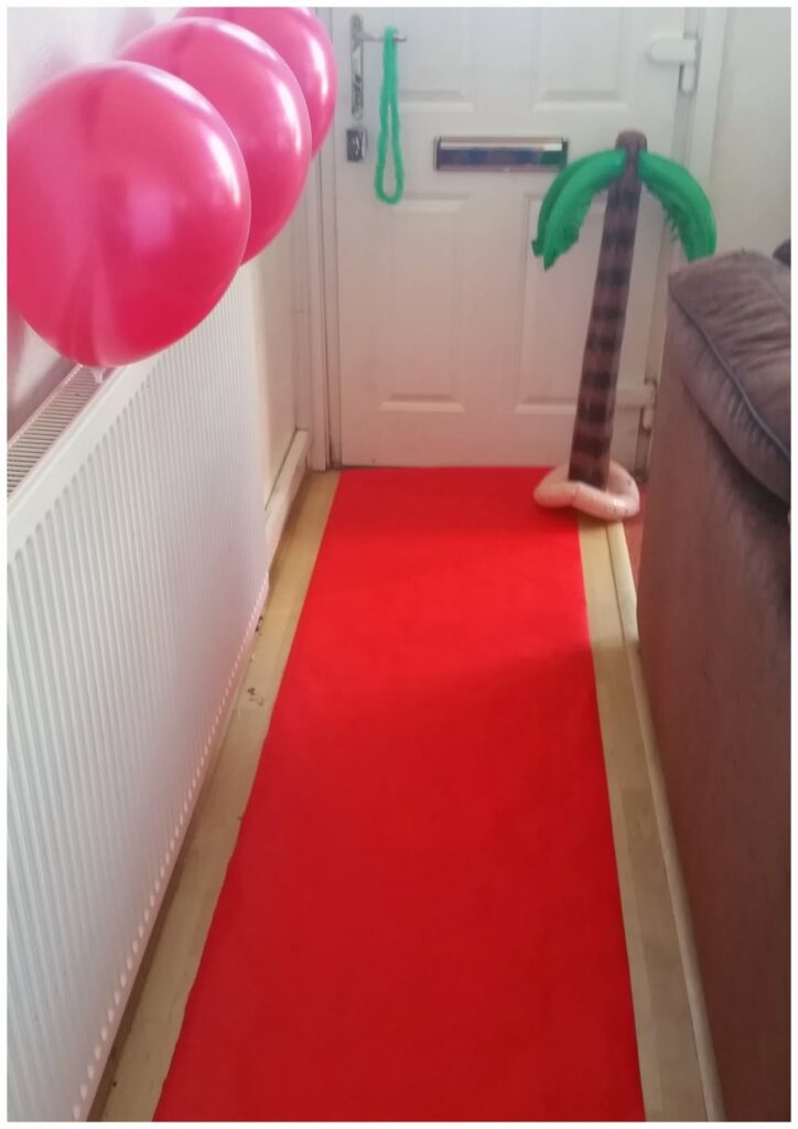 A red carpet runner leads from the foreground to a closed white door. Three red balloons are attached above a radiator on the left. An inflatable palm tree stands on the floor near the door, and part of a brown sofa is visible on the right.