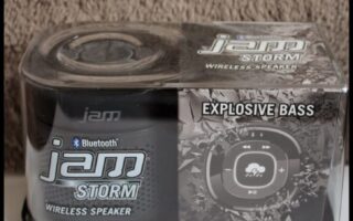 A packaged JAM Storm wireless Bluetooth speaker, labelled “EXPLOSIVE BASS” and “BRING ON THE STORM.” The black and grey box features product branding, a circular speaker graphic, and sits on a white surface against a textured background.