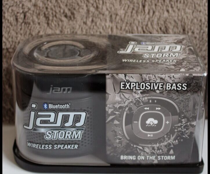 A packaged JAM Storm wireless Bluetooth speaker, labelled “EXPLOSIVE BASS” and “BRING ON THE STORM.” The black and grey box features product branding, a circular speaker graphic, and sits on a white surface against a textured background.