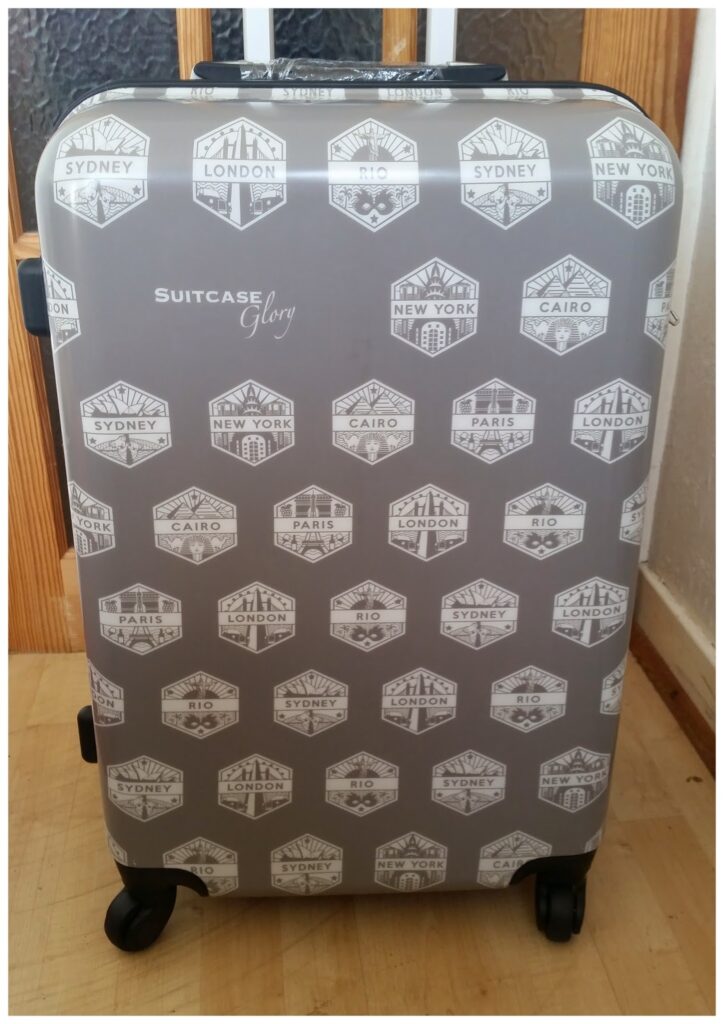 A grey hard-shell suitcase with four wheels is shown standing upright. The suitcase is decorated with white travel-themed badge designs featuring city names like Sydney, London, New York, Paris, Cairo, and Rio. The brand "Suitcase Glory" is printed near the centre.