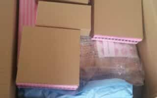 A cardboard box filled with several closed brown and pink boxes, a parcel wrapped in bubble wrap, and some blue tissue paper. The contents appear neatly organised and some items have white labels attached.