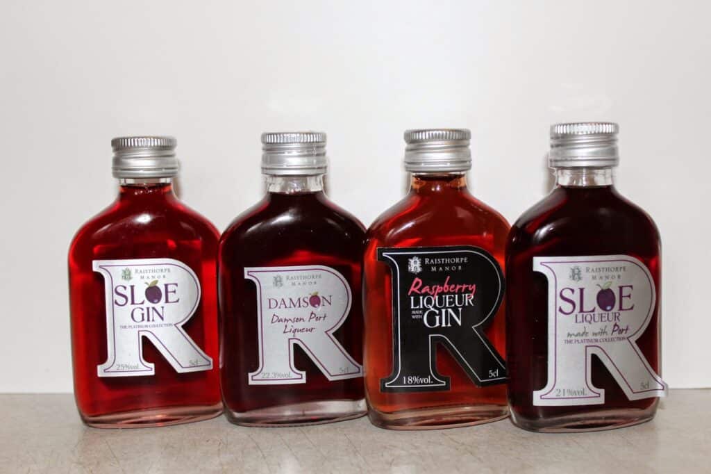Four small glass bottles, each with a silver cap and filled with red or pink liquid, are lined up side by side. The labels read "Sloe Gin," "Damson Port Liqueur," "Raspberry Liqueur Gin," and "Sloe Liqueur." All are from Raisthorpe Manor.