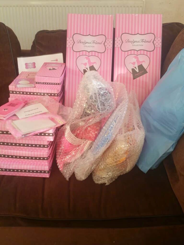 Several pink-and-white striped boxes and two large boxes labelled “DesignaFriend” sit on a brown settee. Three dolls wrapped in bubble wrap and a large blue bag are also on the settee, with a radiator visible in the background.