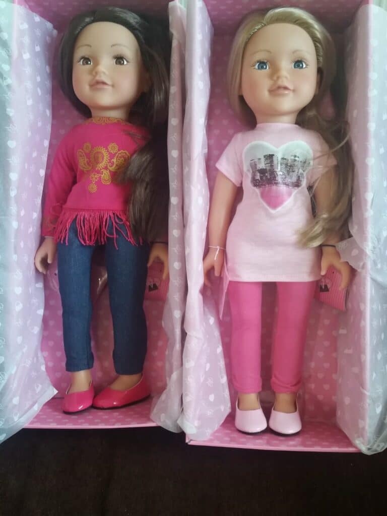 Two dolls in pink heart-patterned boxes. The left doll has long brown hair, a pink fringed top with gold accents, blue jeans, and pink shoes. The right doll has long blonde hair, a pink graphic T-shirt, pink leggings, and light pink shoes.