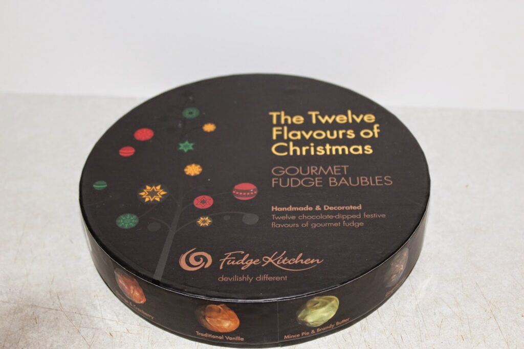 A round black box labelled "The Twelve Flavours of Christmas—Gourmet Fudge Baubles" by Fudge Kitchen. The lid features festive designs and images of fudge. Text mentions handmade, decorated, and chocolate-dipped festive gourmet fudge.