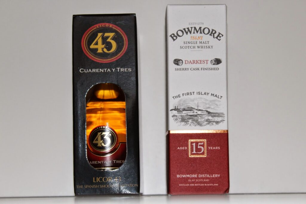 Two boxed bottles stand side by side: on the left, Licor 43 with a black box and gold label; on the right, Bowmore 15 Year Old Single Malt Scotch Whisky in a white box with red and gold accents and an illustration of a distillery.