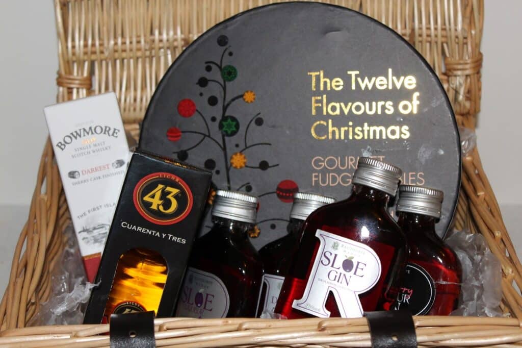 A wicker basket filled with a black tin labelled "The Twelve Flavours of Christmas Gourmet Fudge," small bottles of sloe gin, a box of Bowmore single malt whisky fudge, and a bottle of Licor 43, arranged as a festive gift hamper.