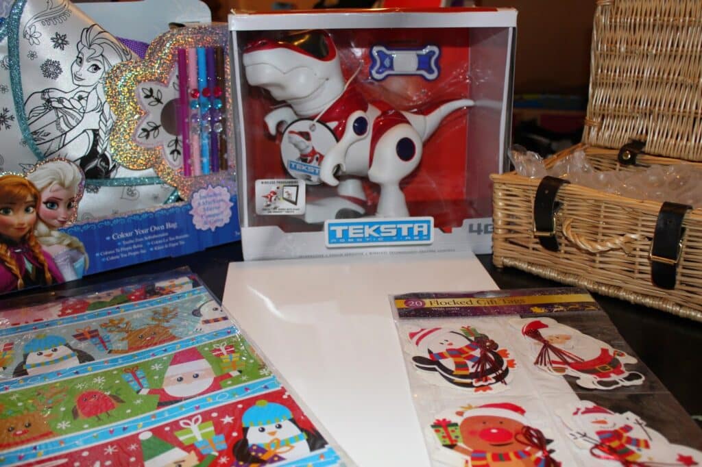 A display of gifts includes a Frozen-themed colouring bag set, a boxed Teksta robot dog toy, a wicker basket, Christmas-themed gift tags with Father Christmas and animals, and festive wrapping paper featuring snowy scenes and holiday characters.