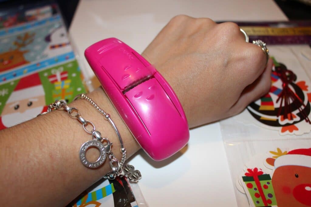 A hand wearing silver bracelets and rings holds a bright pink device that resembles a stapler over the wrist. Christmas-themed papers with images of Father Christmas, reindeer, and baubles are scattered on the table in the background.