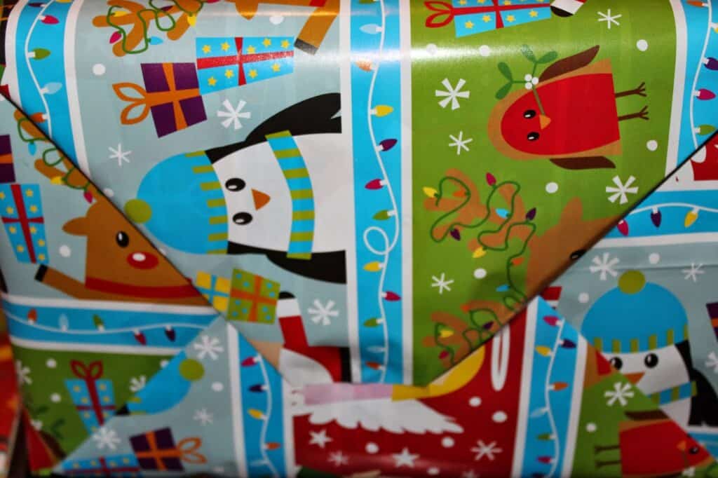 A close-up of a gift wrapped in festive paper featuring penguins in blue hats and scarves, a robin, reindeer, Christmas lights, snowflakes, and colourful presents on a green and blue background. The wrapping paper folds are visible.