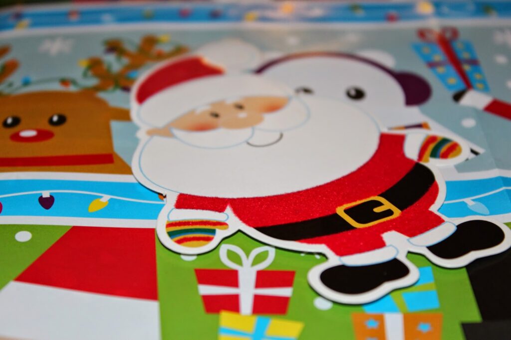 A colourful, cartoon-style Christmas scene featuring a smiling Father Christmas in a red suit, standing among gift boxes. Behind Father Christmas, a reindeer and a snowman appear with holiday decorations and snowflakes in the background.
