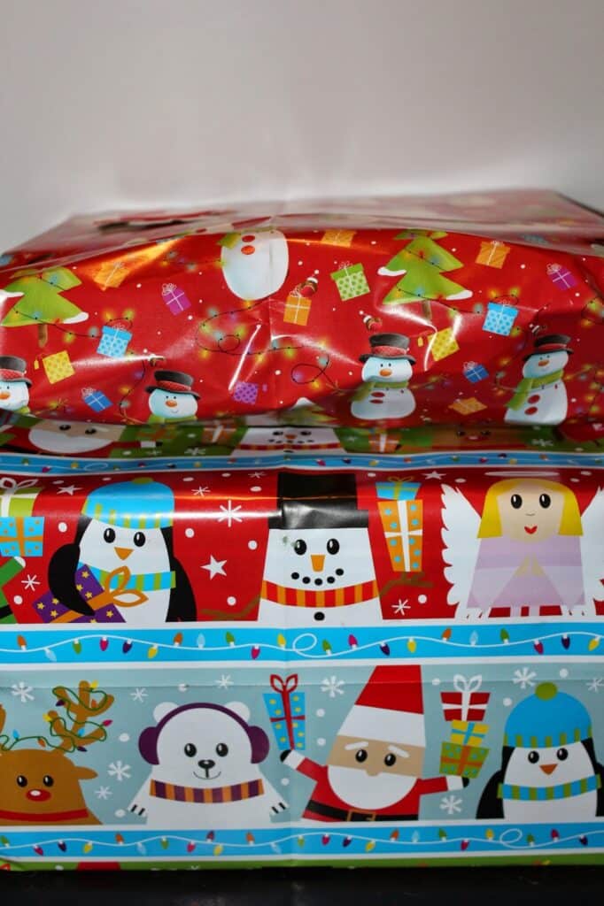 Three stacked gift boxes wrapped in colourful holiday paper. The top box is red with snowmen, Father Christmas faces, trees, and presents. The middle box is blue with penguins and angels. The bottom box has animal faces, Father Christmases, and festive patterns.