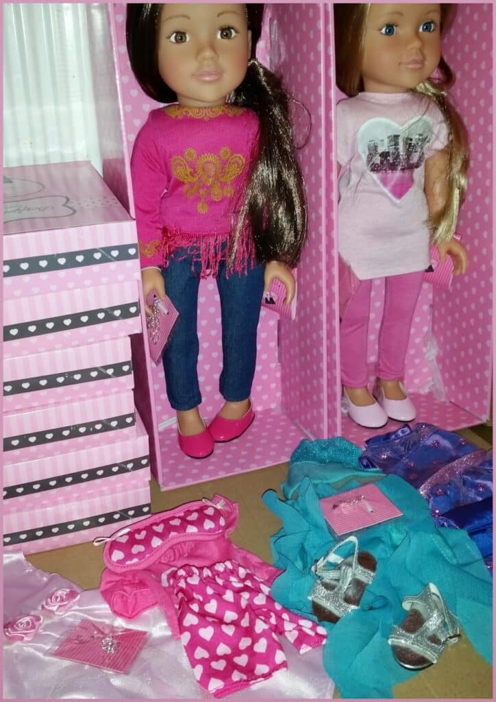 Two dolls with long brown hair stand in pink polka-dot boxes. One wears a pink top and jeans, the other a pink dress and leggings. Around them are doll outfits, shoes, and accessories in pink, teal, and silver, displayed neatly on a surface.