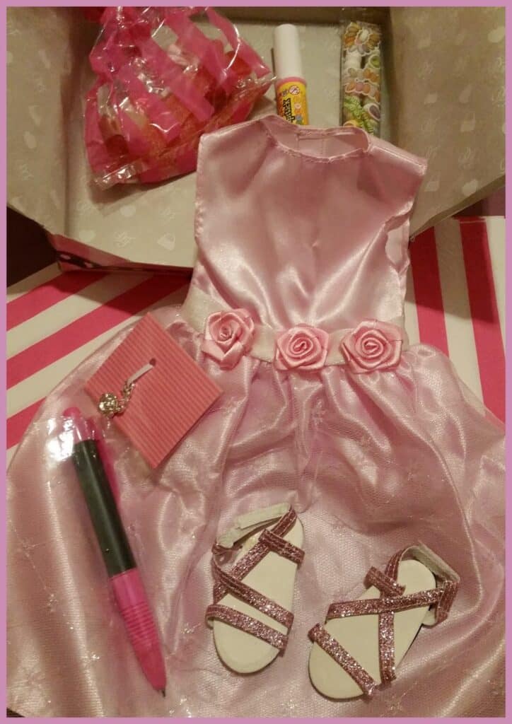 A shiny pink dress with rose accents, glittery pink sandals, a pink pen, a small pink notebook, a tube of lip balm, and a bag of star-shaped hair clips are arranged in an open box with a white heart pattern inside.