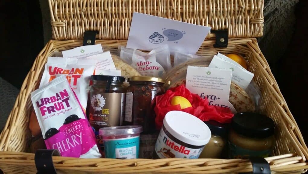 A wicker hamper filled with assorted treats, including jars of Nutella, honey, and preserves, packets of Urban Fruit snacks, cheese, wraps, fruit, and various packaged goods. A greeting card rests at the back inside the open lid.