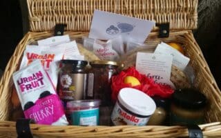A wicker hamper filled with assorted treats, including jars of Nutella, honey, and preserves, packets of Urban Fruit snacks, cheese, wraps, fruit, and various packaged goods. A greeting card rests at the back inside the open lid.